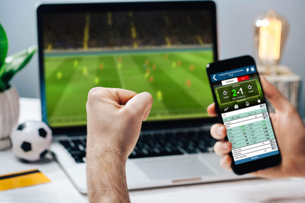 obrazek przedstawia zaciśniętą pięść oraz smartfon z typami na żywo, a w tle mecz na laptopie