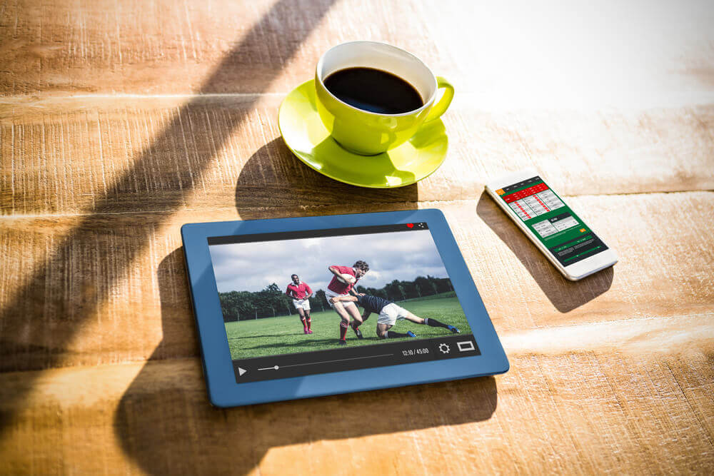 Obrazek przedstawia filiżankę kawy, tablet z meczem futbolu oraz smartfon z aplikacją do obstawiania spotkań