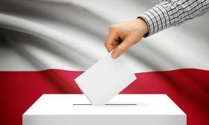 obrazek przedstawia rękę wrzucającą głos do urny wyborczej, na tle polskiej flagi